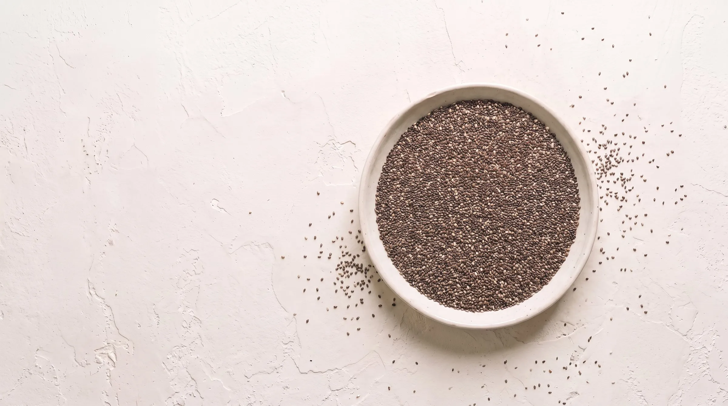 A shallow pale ceramic dish of dry chia seeds seen from directly above on warm off-white plaster, the seeds tiny mottled grey, brown and cream ovals, with loose seed scattered on the plaster around the dish.