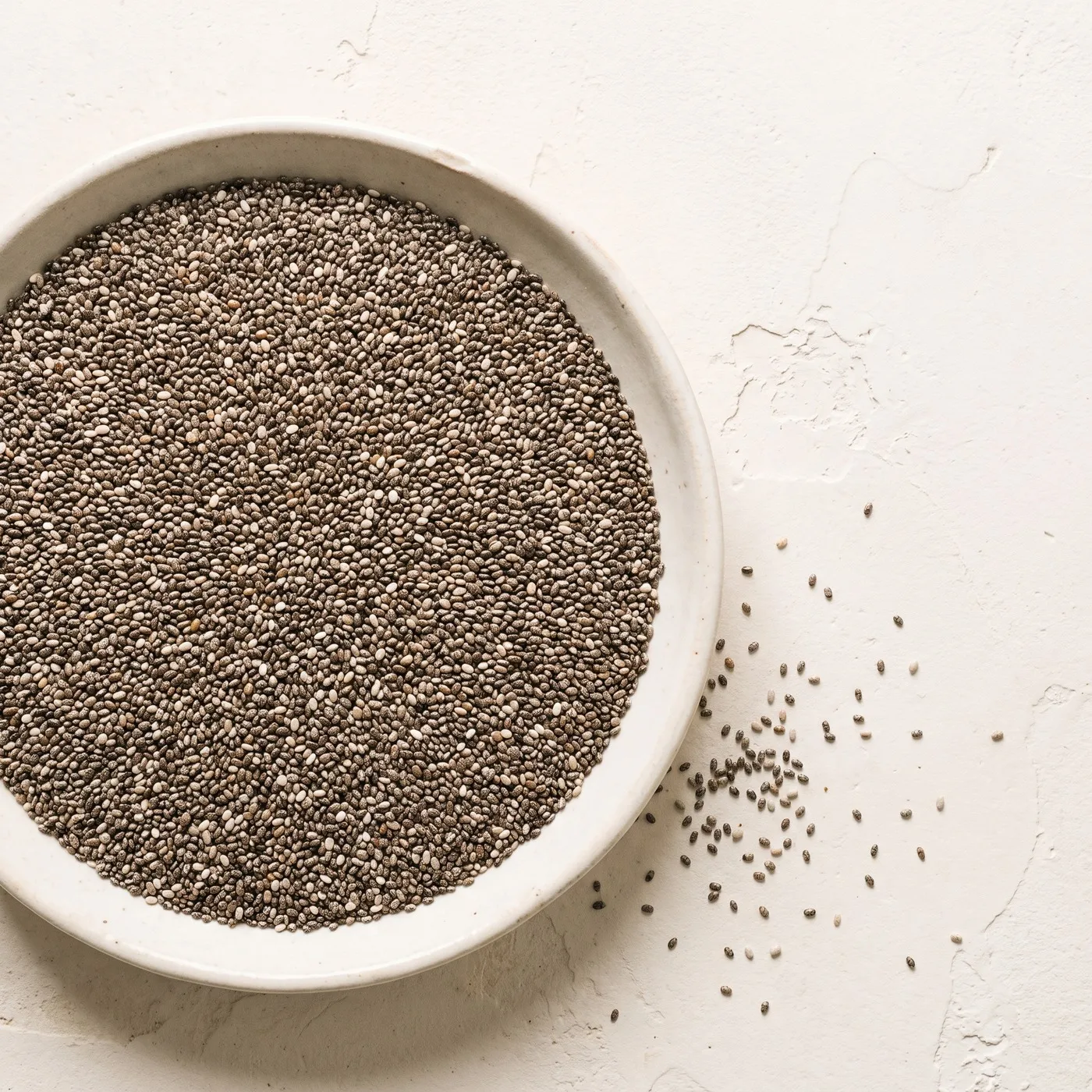 A shallow pale ceramic dish of dry chia seeds seen from directly above on a plain warm off-white surface, the seeds tiny mottled grey, brown and cream ovals, with a light scatter of loose seed on the surface beside the dish.